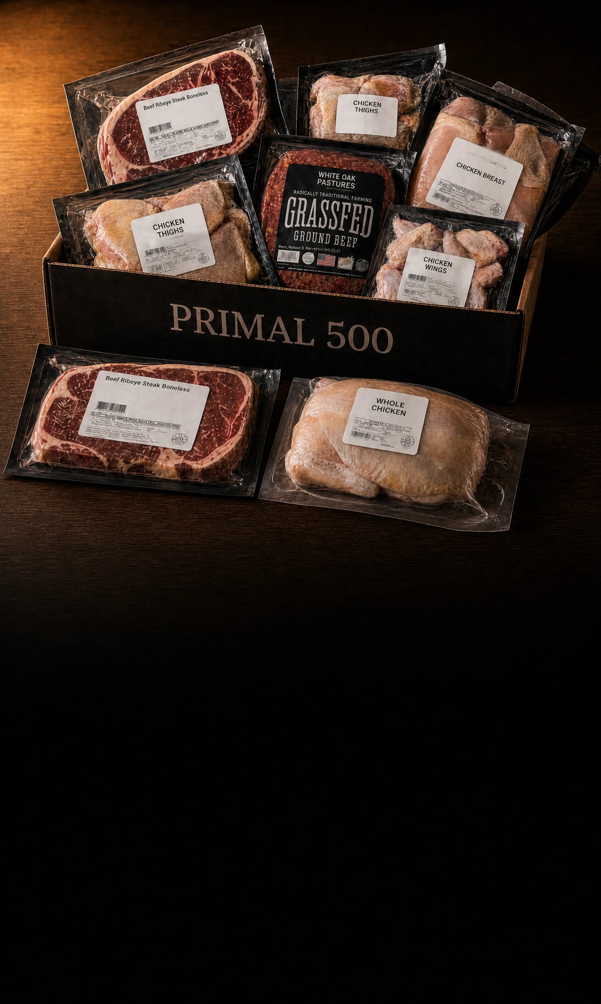 The Primal 500 Founding Member Box on a wooden farmhouse table, packed with labeled vacuum packs: ribeye steaks, White Oak Pastures grass-fed ground beef, chicken breast, thighs and wings, with a second ribeye and a whole chicken resting on the table in front of the box.